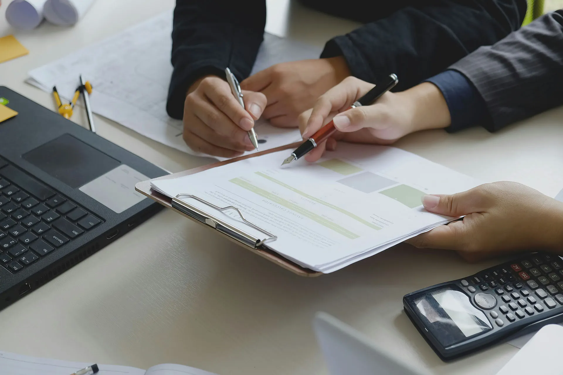  Two people point pens at a document on a clipboard during a business meeting, with a laptop and a calculator visible on the desk.