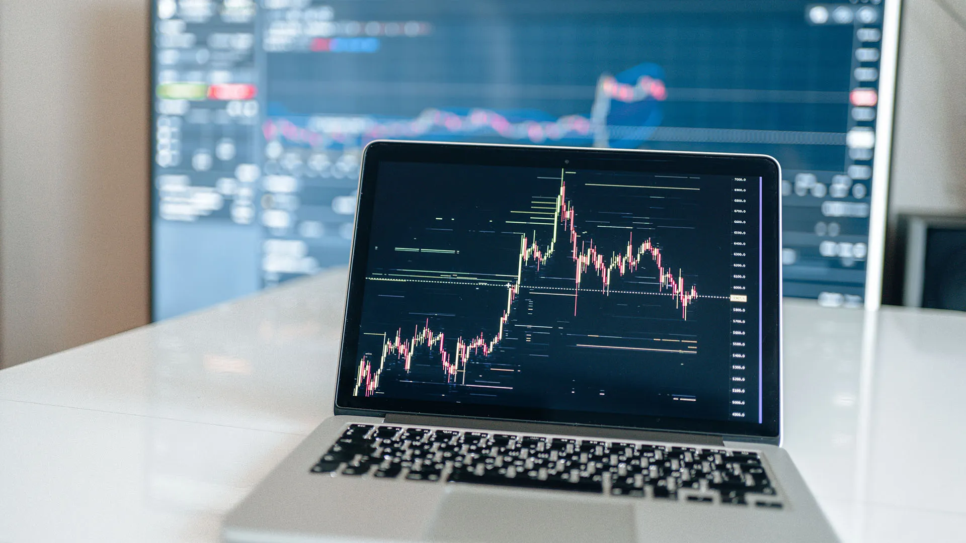 A hand points a pencil at printed financial charts on a desk next to a smartphone calculator, with a laptop in the foreground and a large monitor displaying stock market candlestick charts in the background.