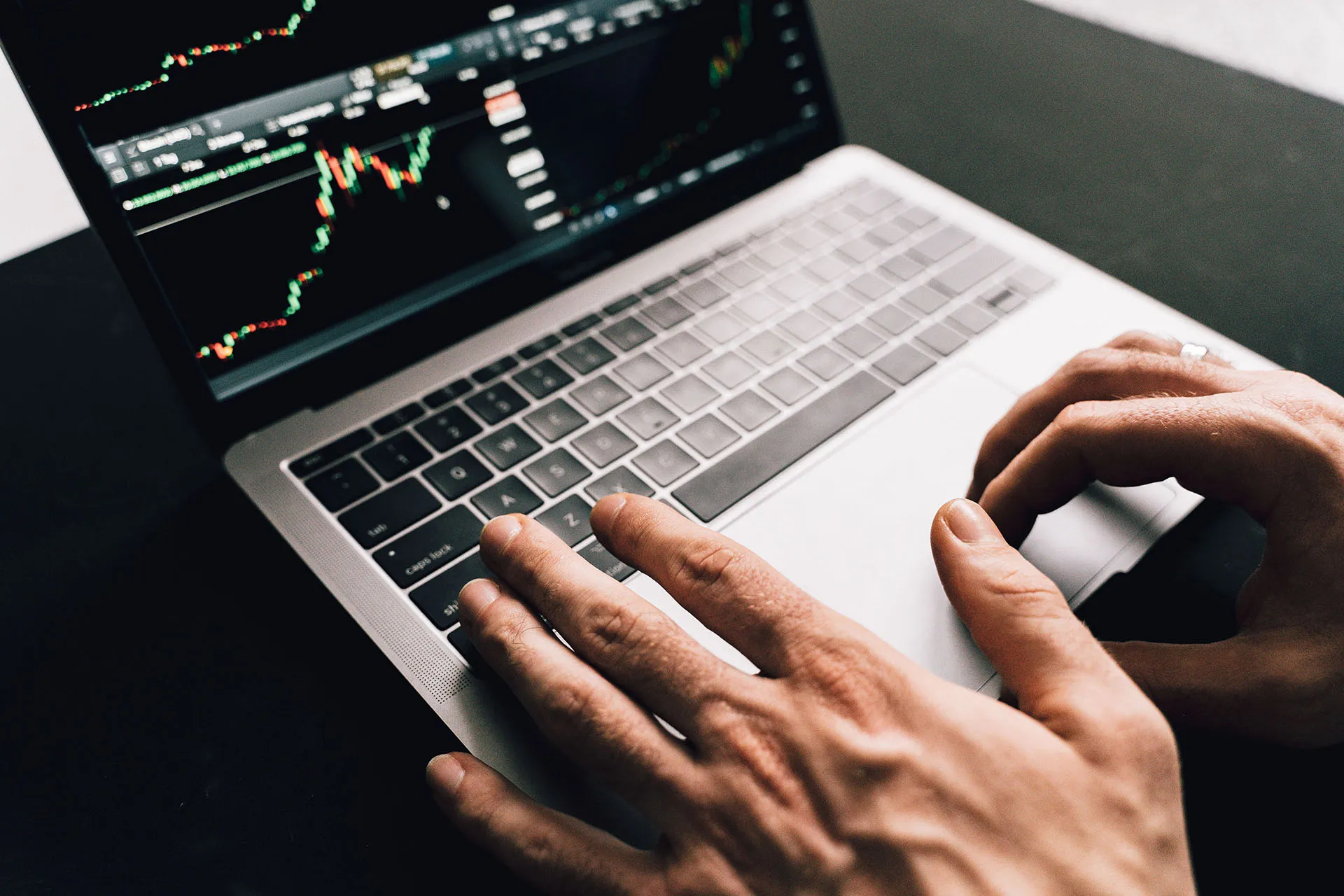  An overhead view of a user's hands typing on a silver laptop keyboard while analyzing live market trading charts on the screen.