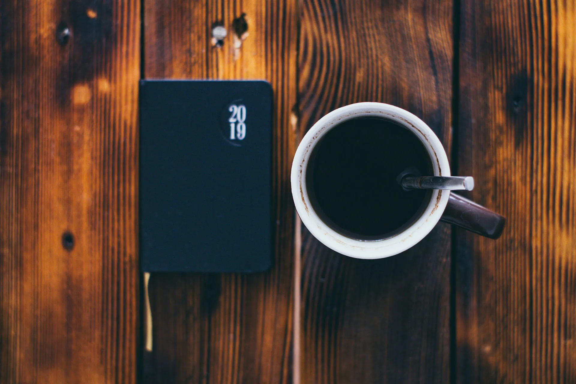  Black notebook that says 2019 in white text on a wooden table beside a coffee mug