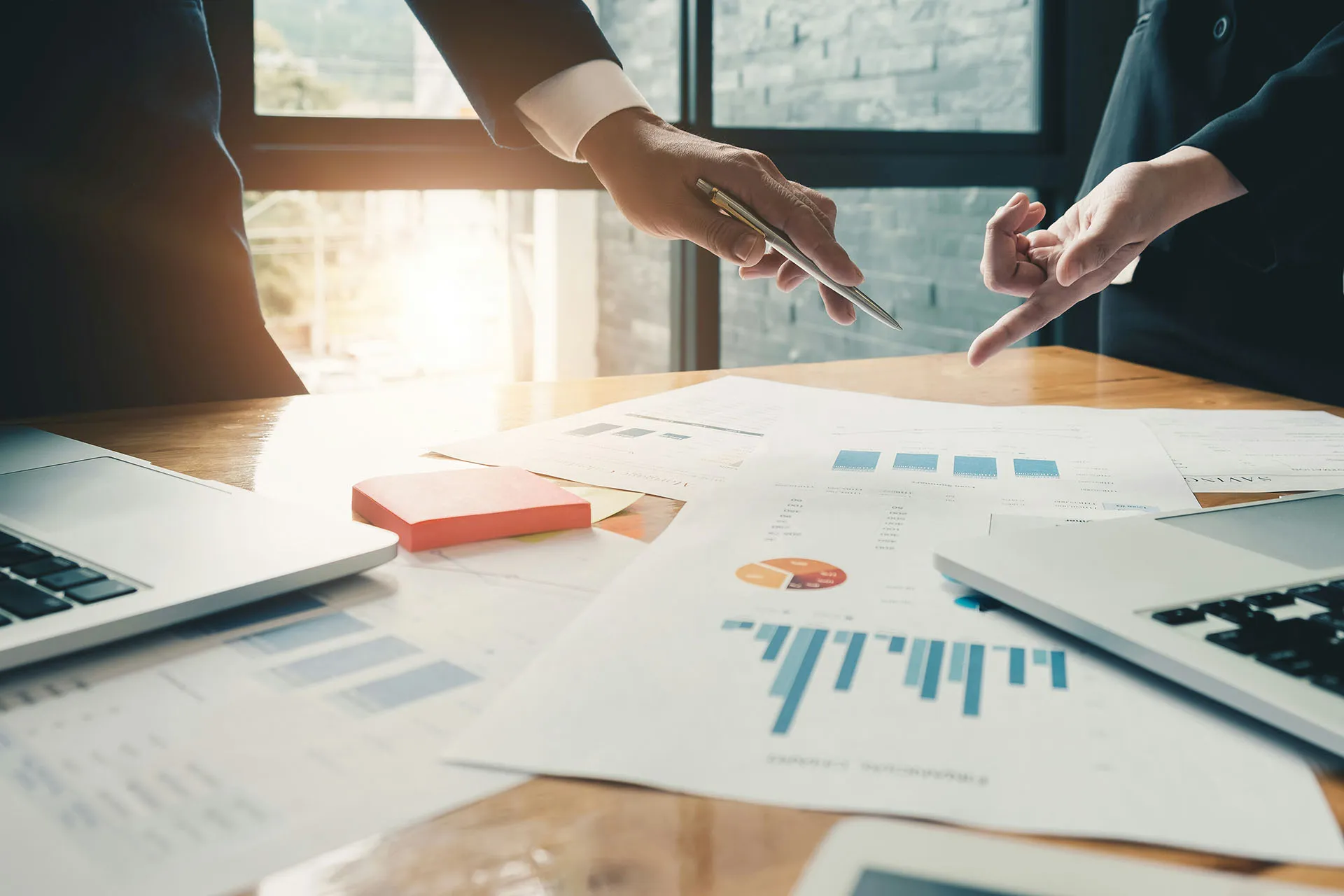  A close-up shot of financial colleagues pointing at data charts, bar graphs, and performance metrics on a conference table during a portfolio strategy review.