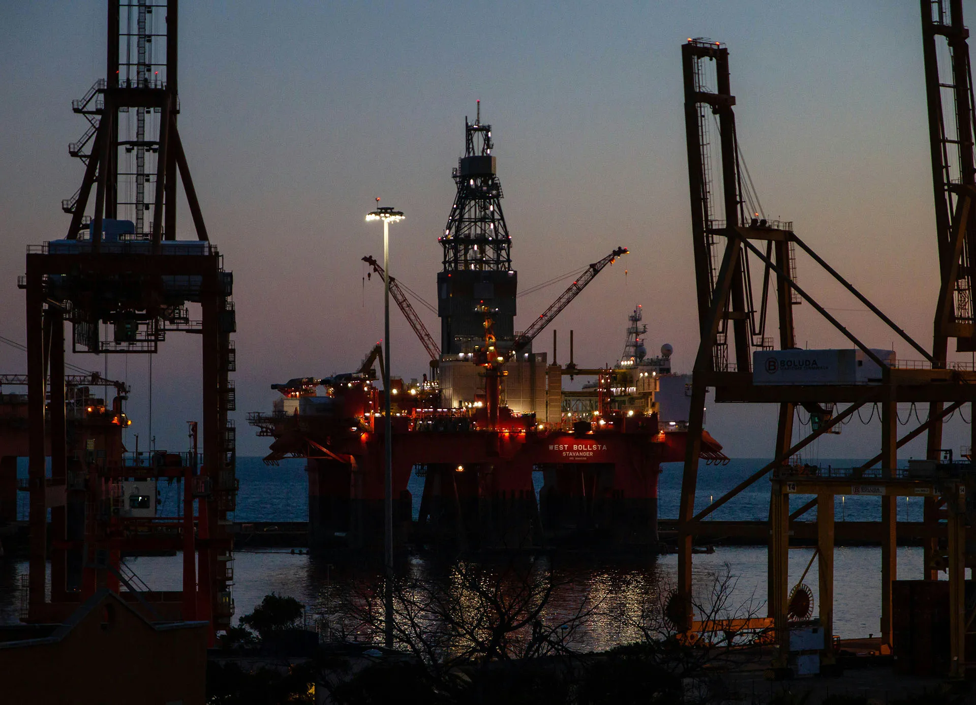 Offshore oil rig and industrial cranes at dusk, with lights reflecting on the water.