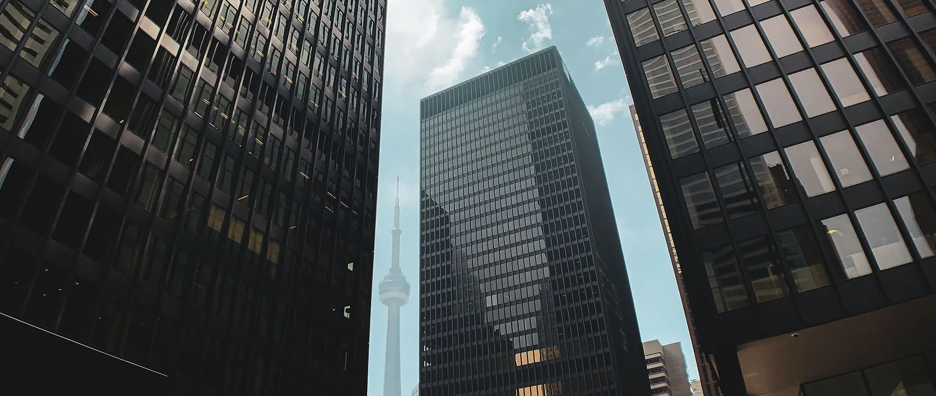  A low-angle view of the CN Tower rising above modern city skyscrapers and office buildings under a blue sky with clouds.