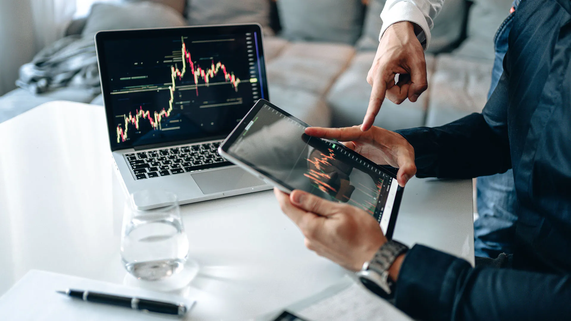  Business professionals gesturing to a digital tablet displaying an upward-trending stock market candlestick chart next to a laptop with analytical financial graphs.