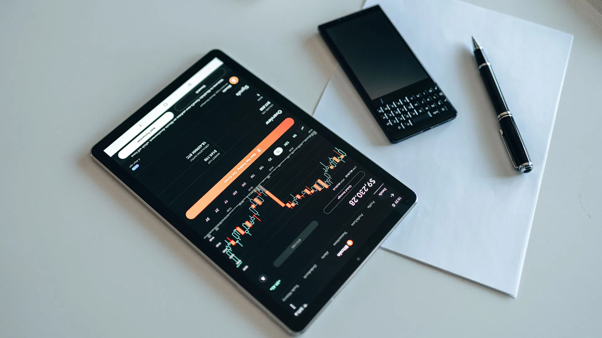 A tablet displaying fluctuating financial charts beside a blank piece of paper, a pen and a smartphone all on a white table
