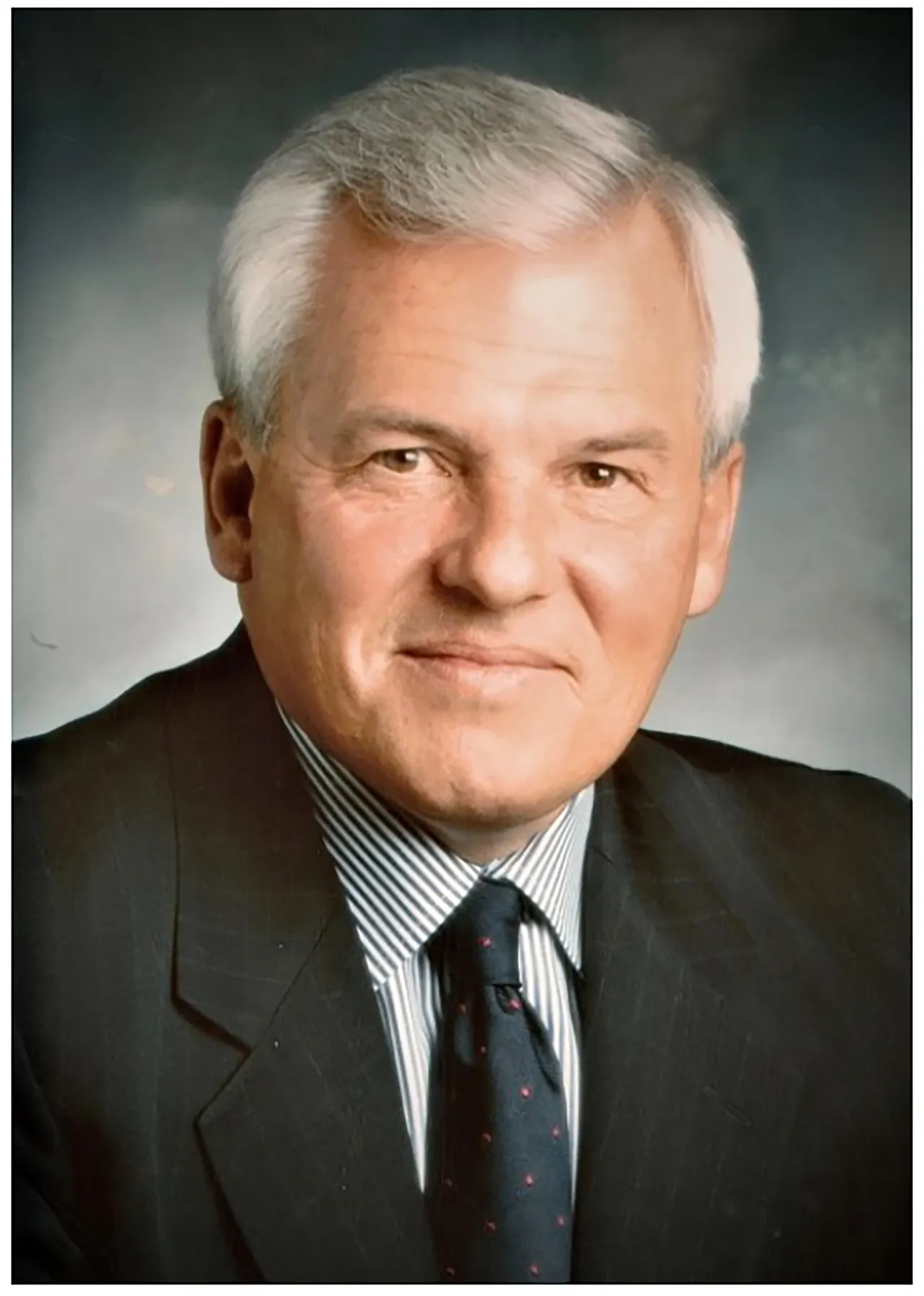 Professional headshot of an older man with white hair wearing a dark suit and tie against a neutral studio background.