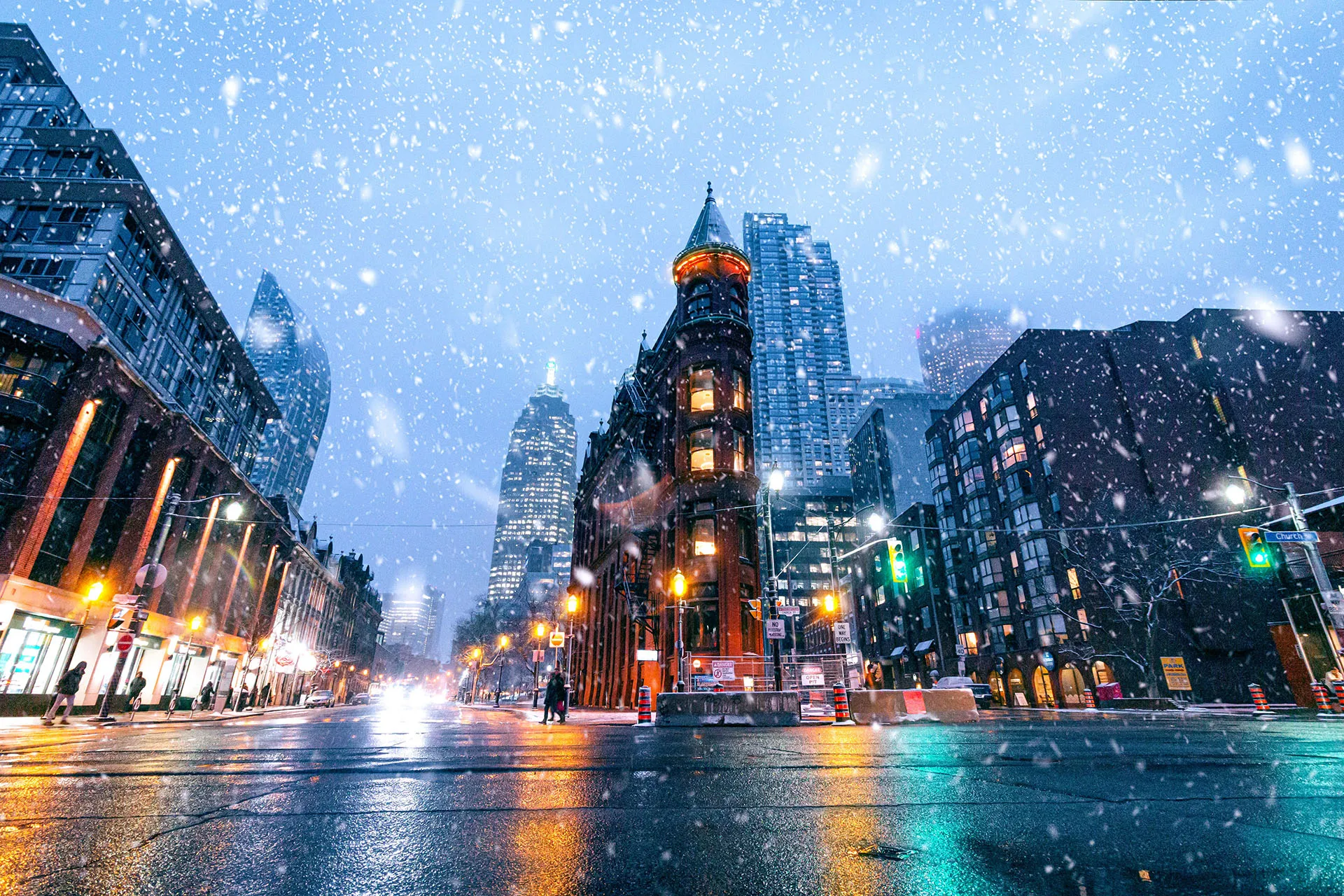  A street-level view of a road at night during snowfall, centered on an illuminated building in downtown Toronto flanked by lit skyscrapers and city traffic.