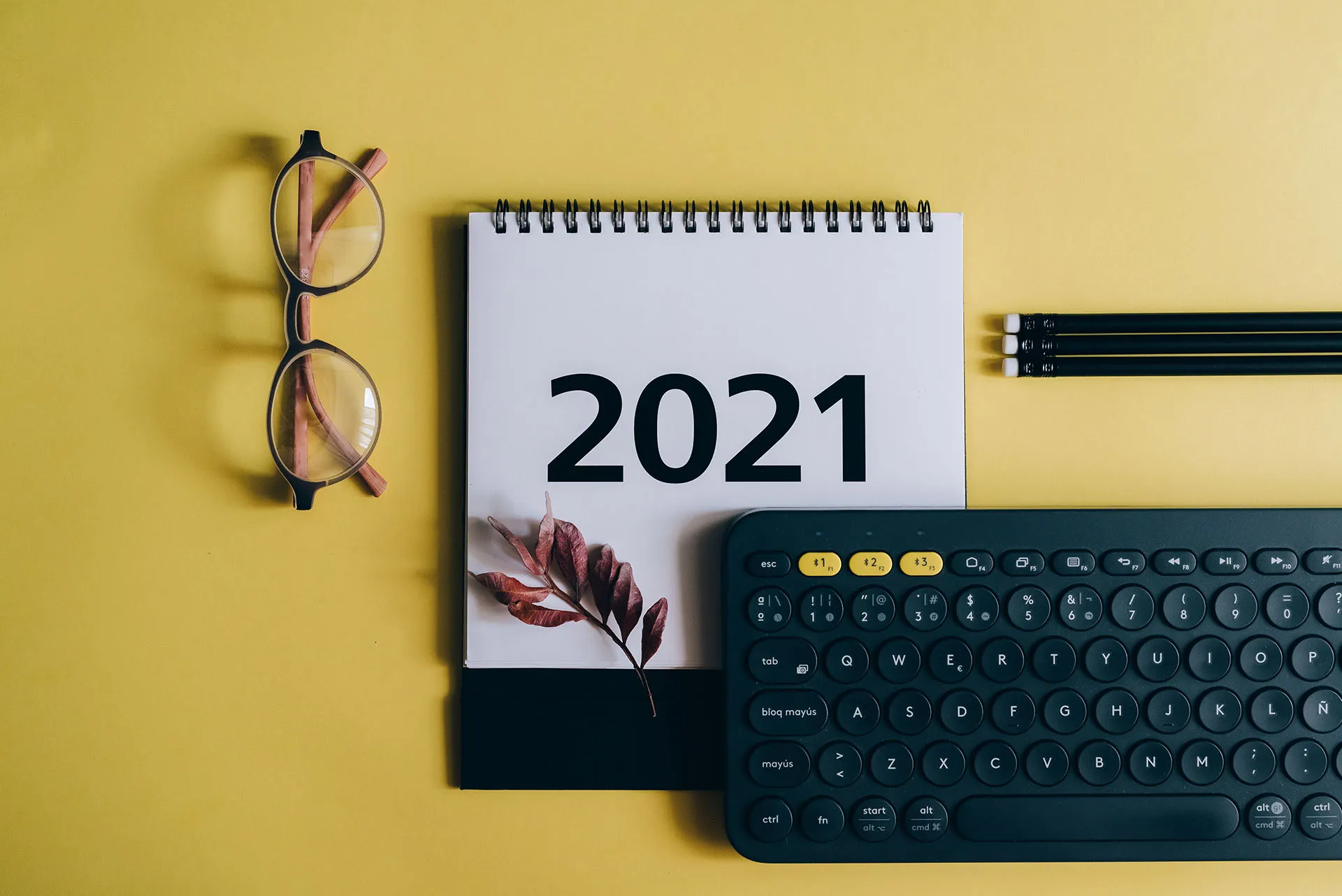  White desk calendar showing the year 2021 beside a computer keyboard, pencils, and a pair of glasses on a yellow table.