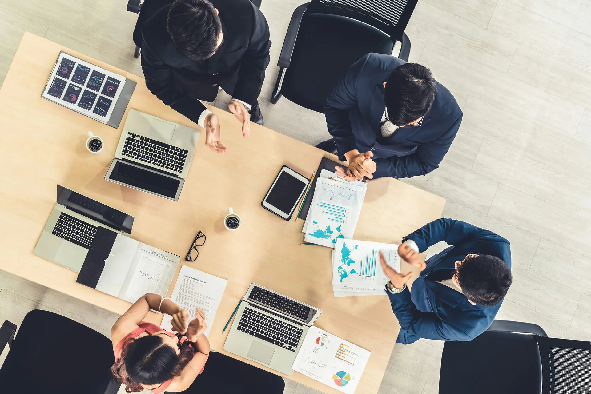  An overhead view of a corporate business team in suits sitting around a conference table filled with laptops, financial reports, and charts, clapping during a meeting.