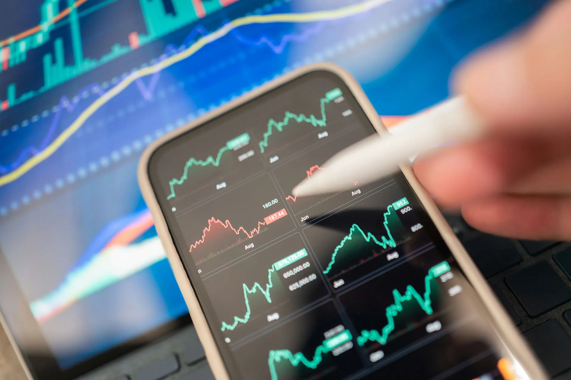  A close-up shot of a person using a stylus on a smartphone screen displaying multiple volatile financial stock charts, with a larger market dashboard blurred in the background.