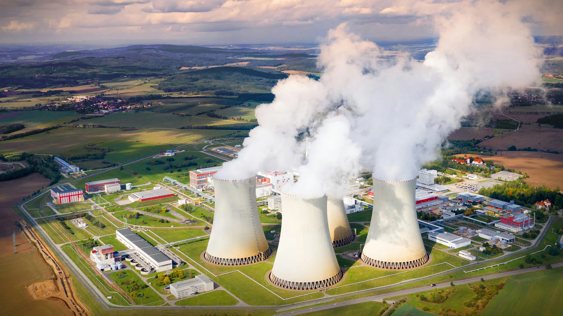  Aerial view of a nuclear power plant with cooling towers releasing steam over a rural landscape.
