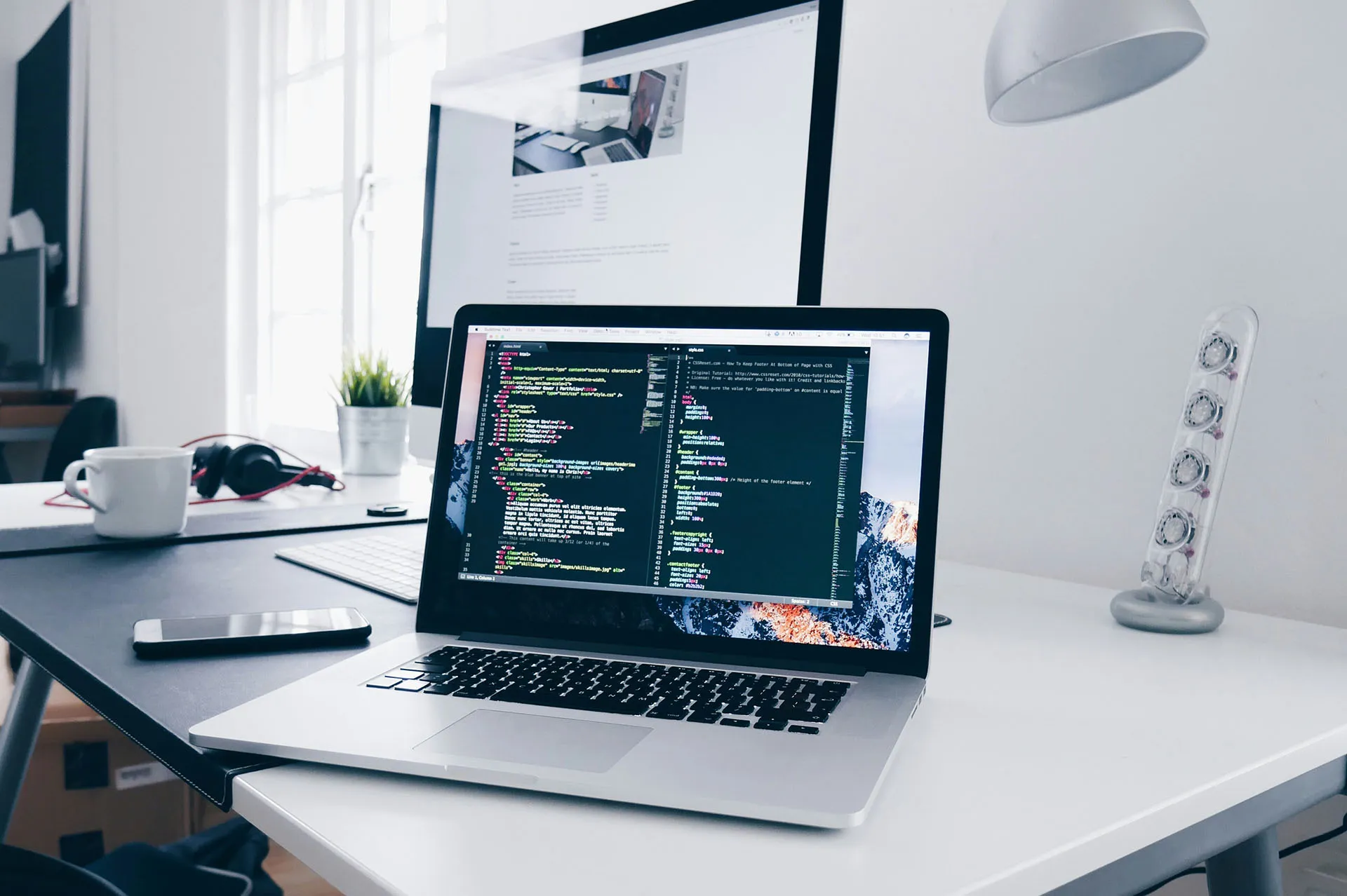  Laptop displaying code on a clean office desk with a monitor, keyboard, headphones, and workspace accessories.