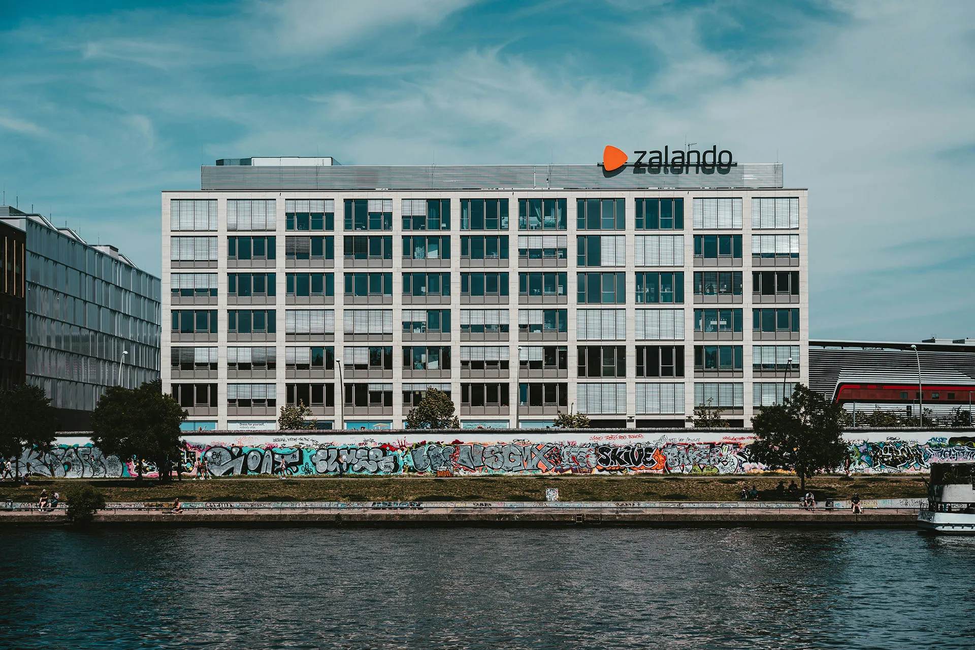  Zalando office building along a waterfront with graffiti-covered wall and cloudy blue sky.