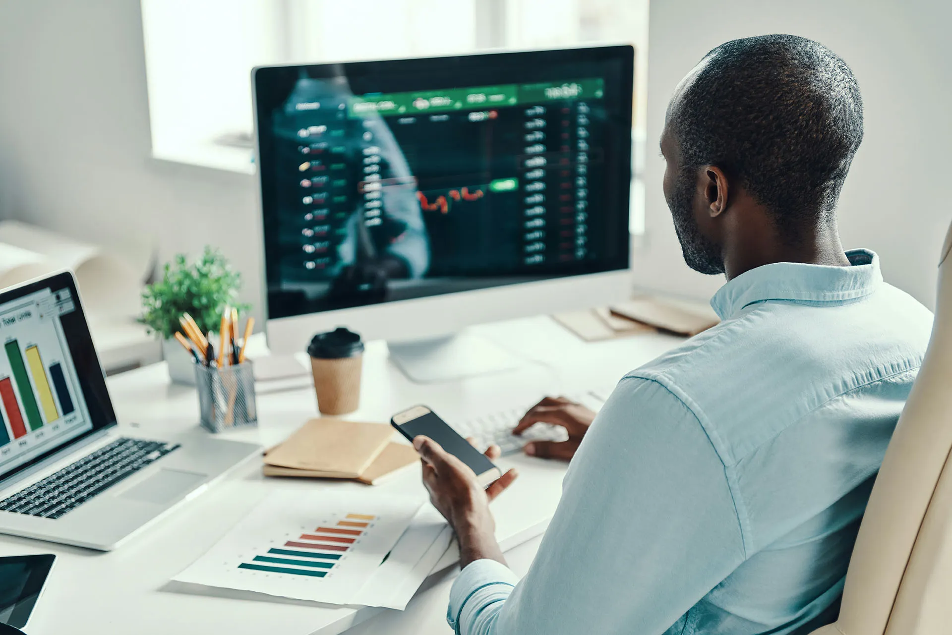  An over-the-shoulder view of a financial professional sitting at a desk, looking at a large computer monitor displaying a stock trading dashboard while holding a smartphone.