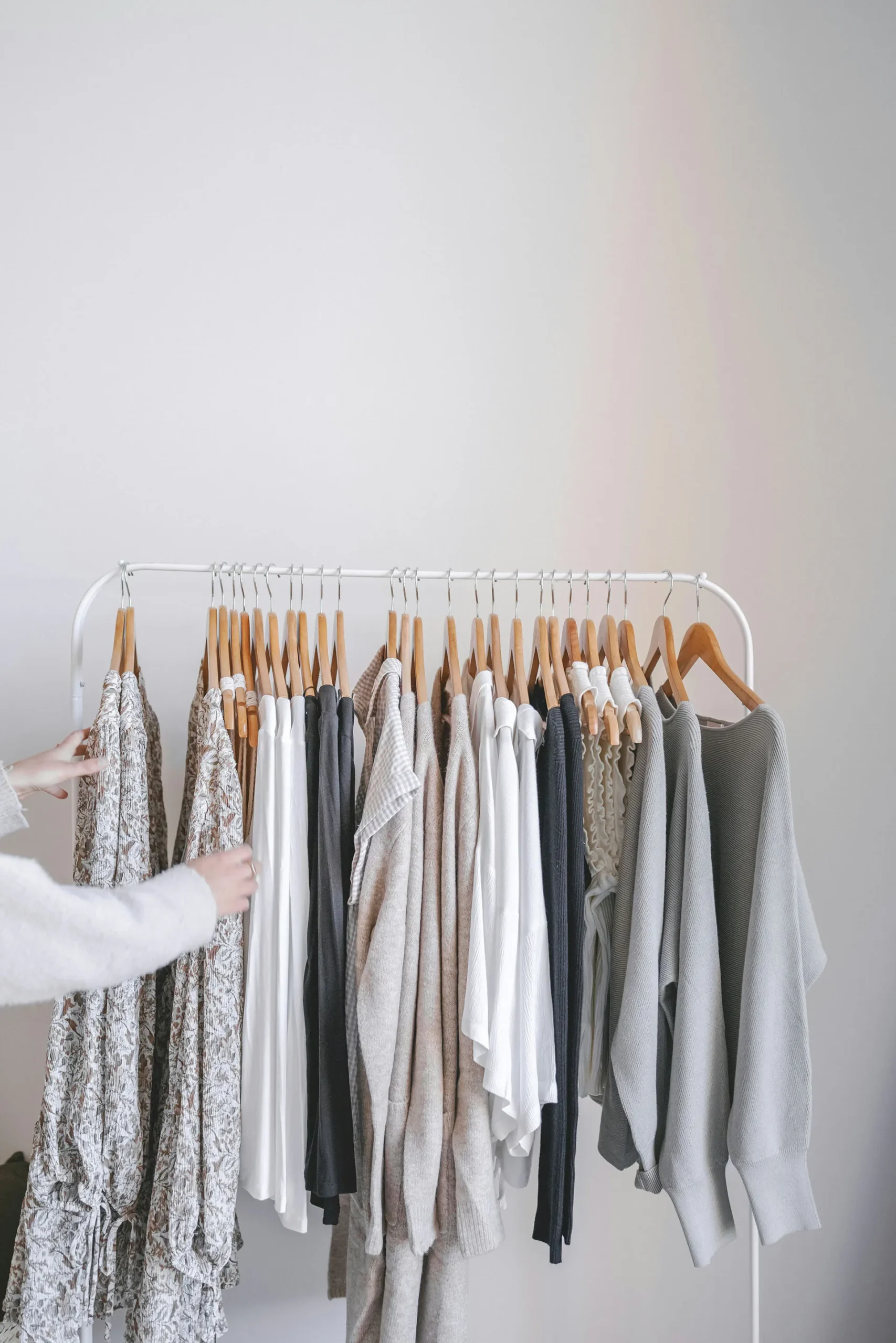 Person browsing a neutral-toned clothing rack with dresses, sweaters, and shirts on wooden hangers.