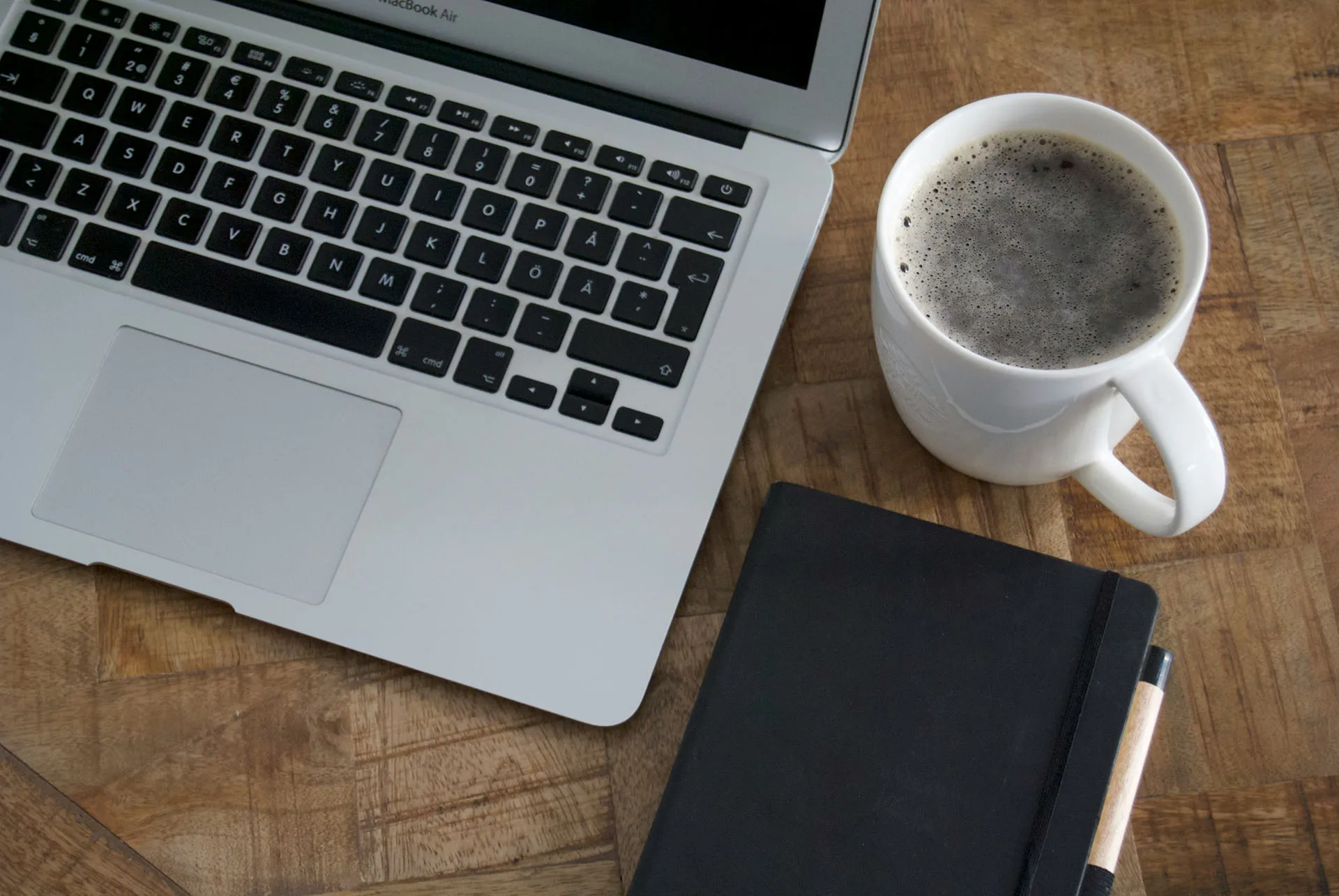  An overhead view of a workspace on a wooden desk featuring an open laptop, a white mug filled with black coffee, and a black hardcover notebook with a pen.