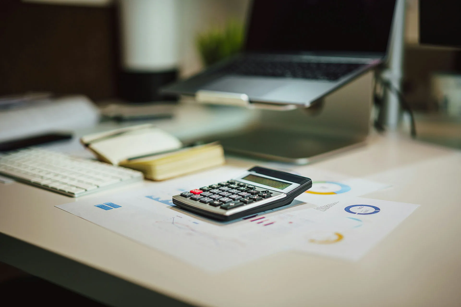 Calculator on financial charts at an office desk with laptop, keyboard, and notebook in the background.