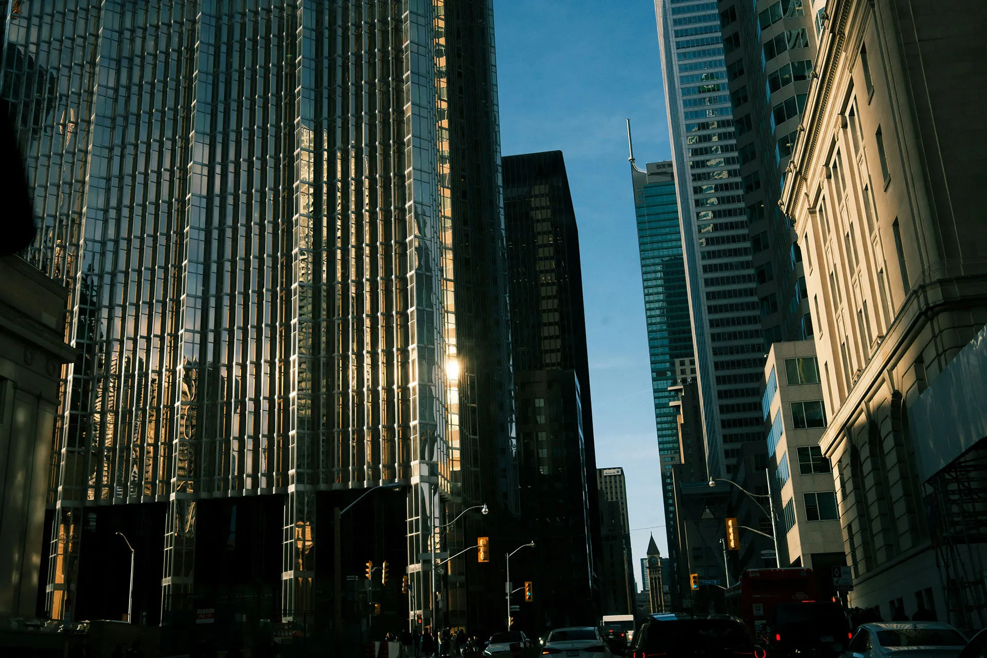  A low-angle view looking down a busy financial district street in Toronto, flanked by towering modern skyscrapers reflecting the afternoon sunlight under a clear blue sky.
