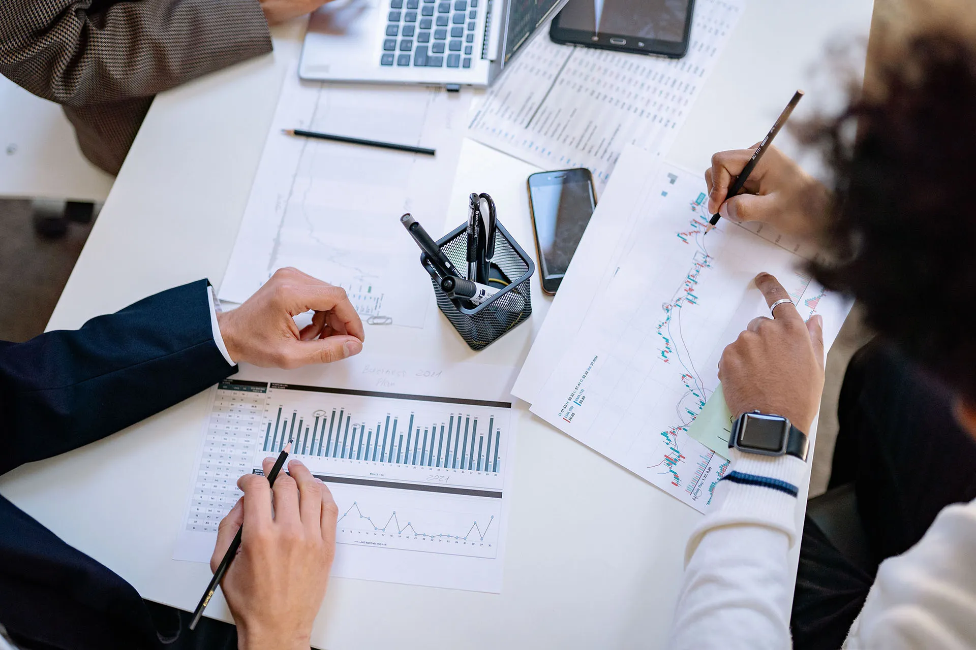 An aerial view of financial professionals around a table at a business meeting, writing on stock charts and typing on laptops