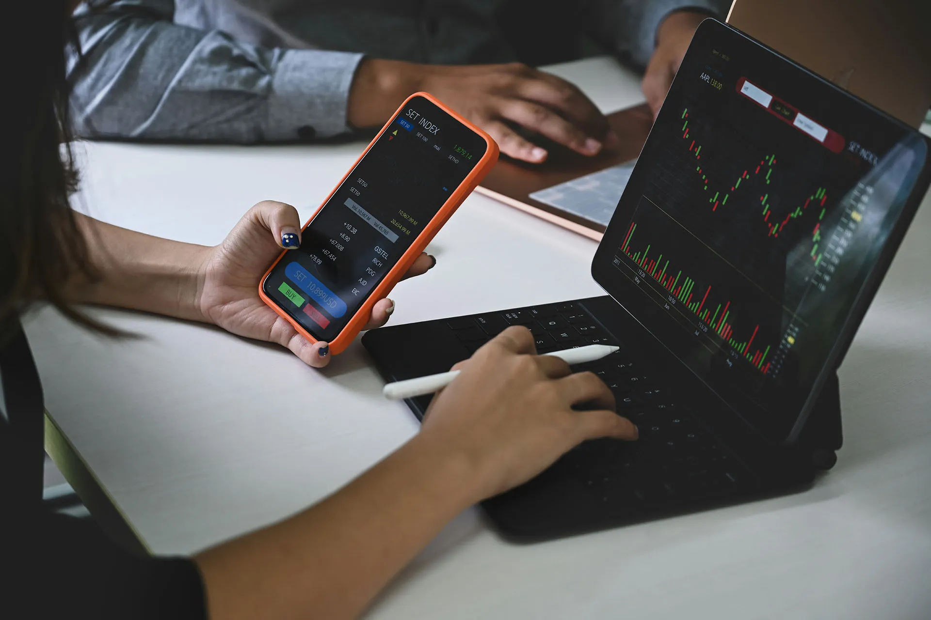  A financial professional uses a smartphone and a tablet to track stock market data and candlestick charts at a desk.