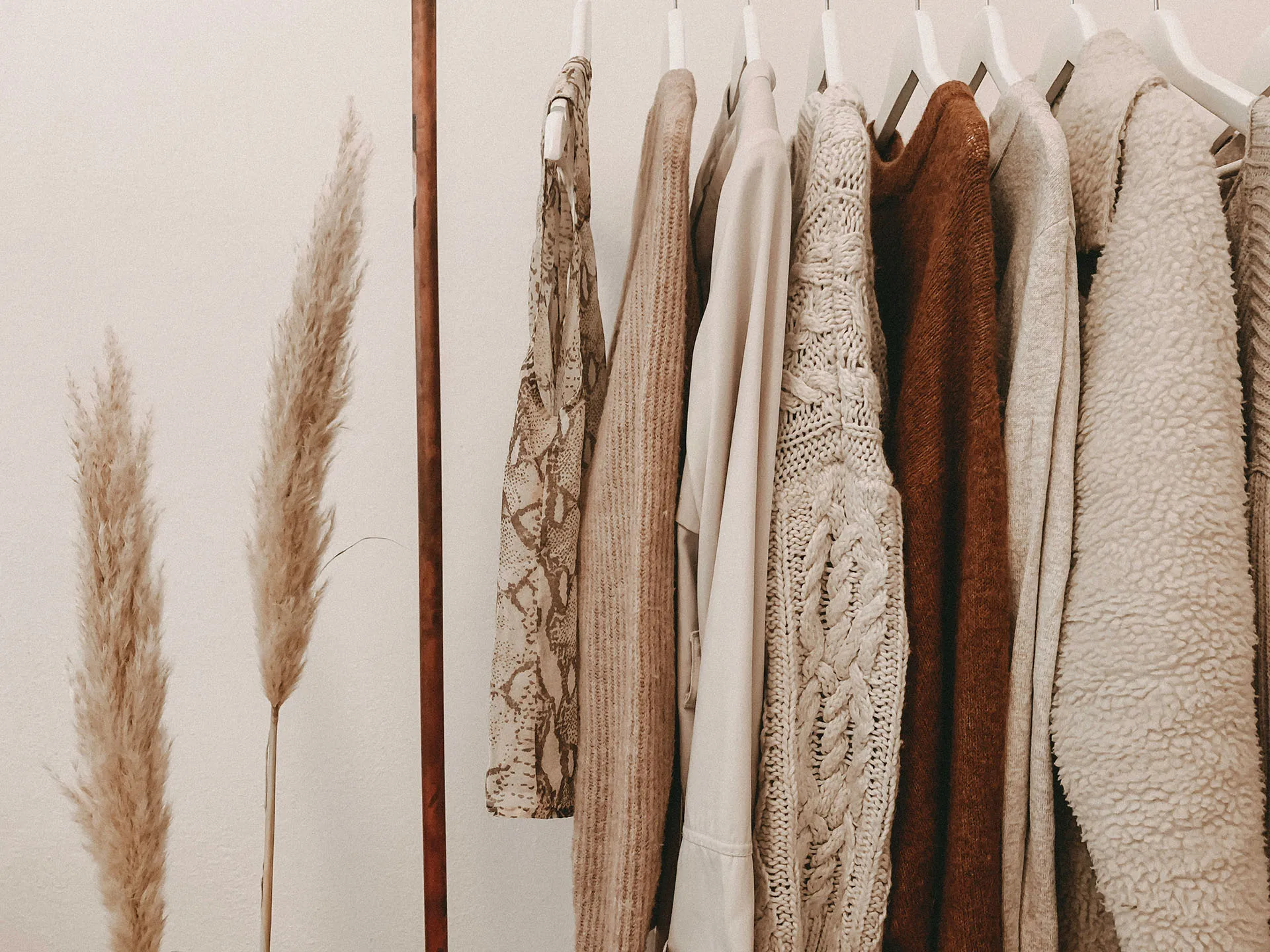  Neutral clothing rack with textured sweaters, coats, and pampas grass in a minimalist boutique setting.