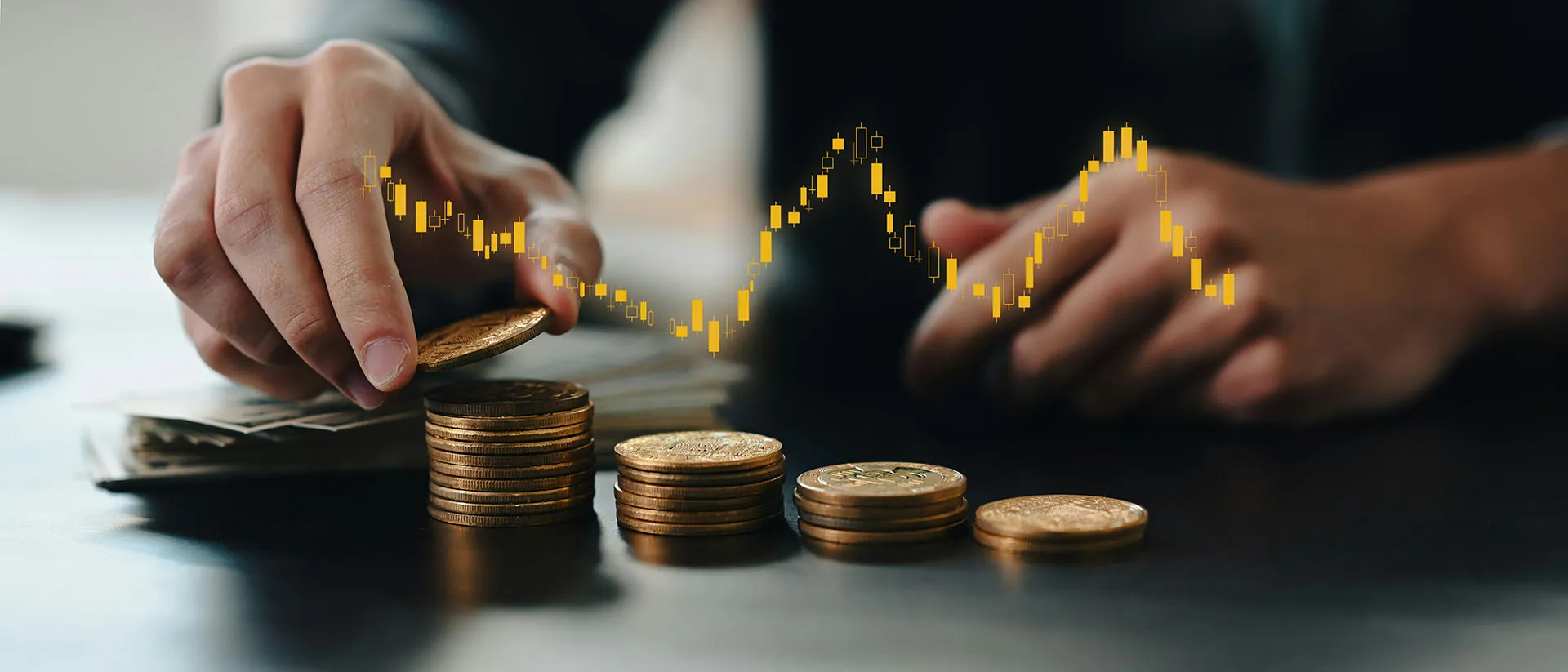  An investor's hands stacking gold coins on a desk alongside a digital overlay of a fluctuating golden candlestick market chart.