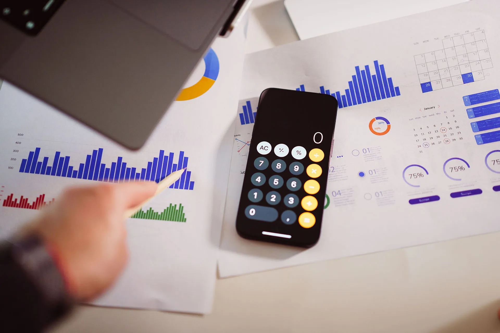  Calculator on financial charts at a desk with a laptop, keyboard, and a person's hand gesturing towards them with a pencil.