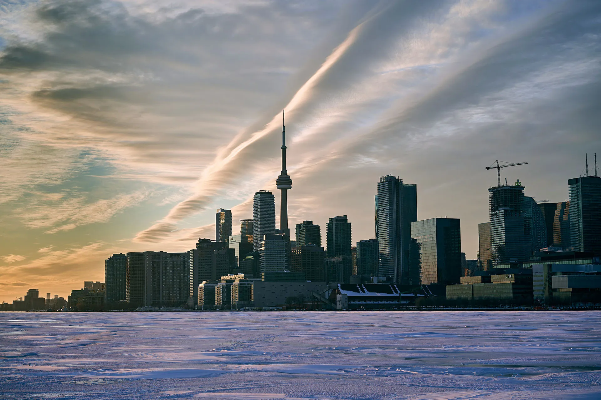  A wide shot of the Toronto city skyline in the winter, featuring the CN Tower, under a dramatic, cloudy sunset sky with a frozen lake in the foreground.