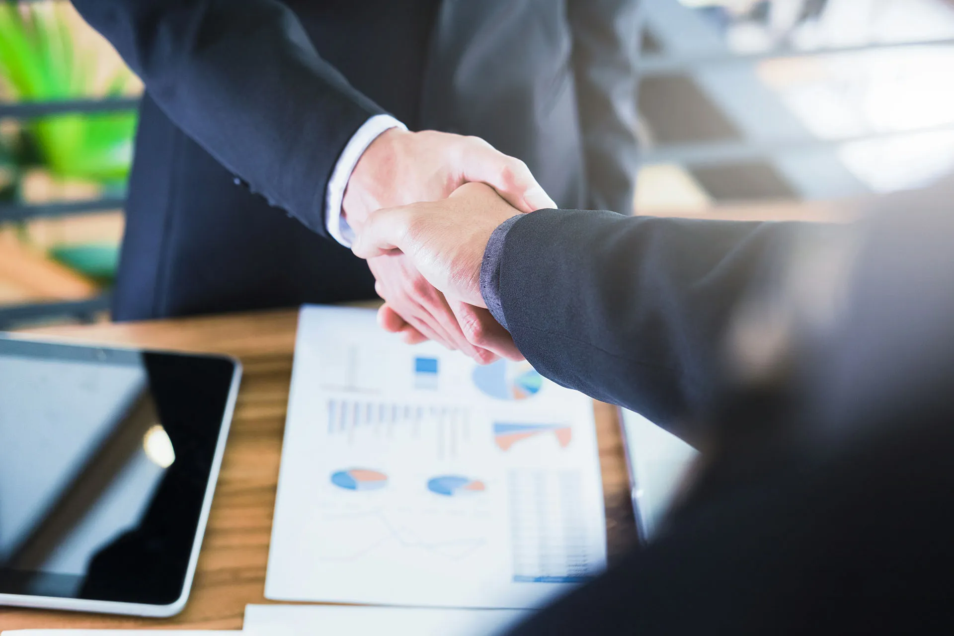  Two business professionals in suits shaking hands over a wooden office desk featuring financial data charts and a tablet.