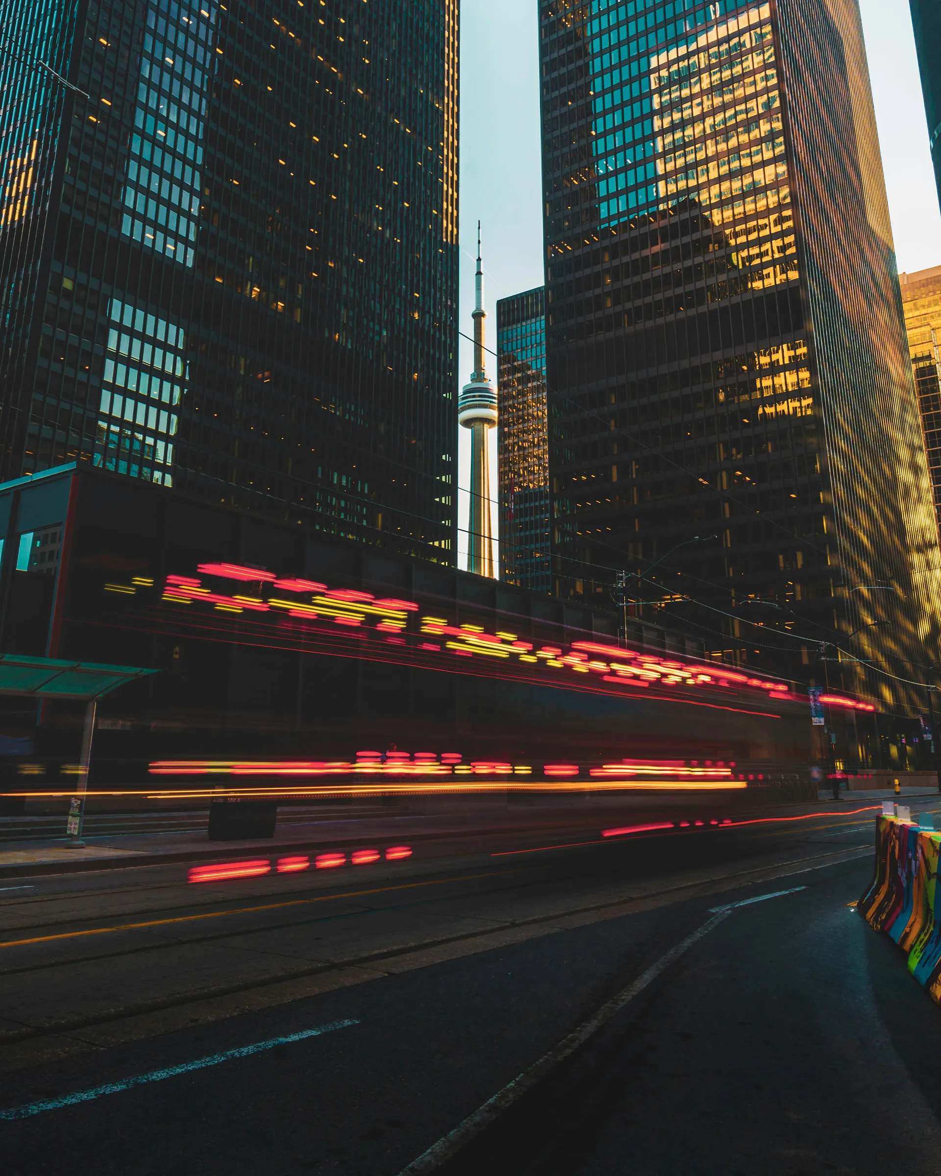  A long-exposure shot of a city street with red and yellow light trails from moving traffic, framed by towering skyscrapers and the CN Tower in the background.