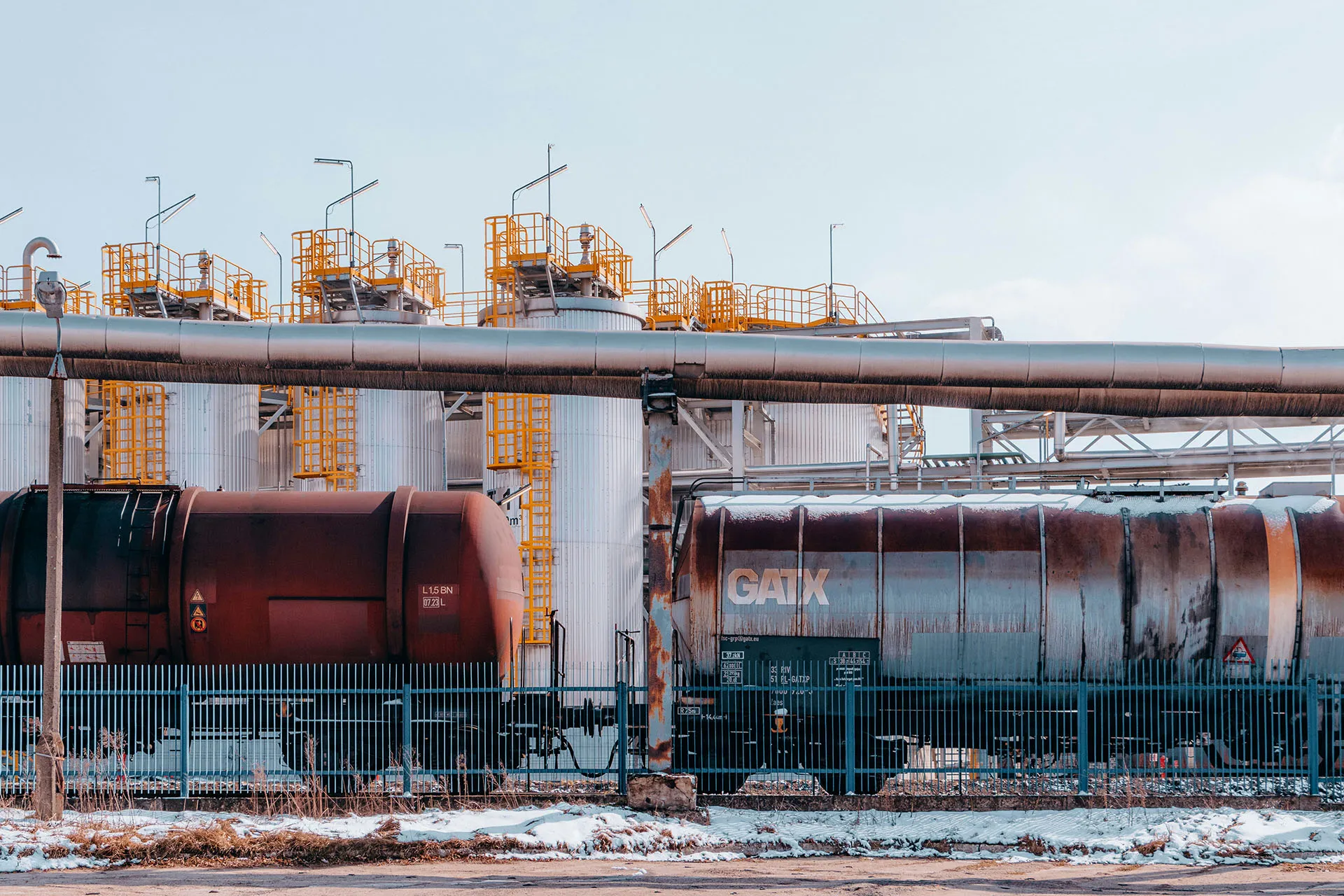  Industrial facility with storage tanks, pipelines, and rail tank cars behind a fence.