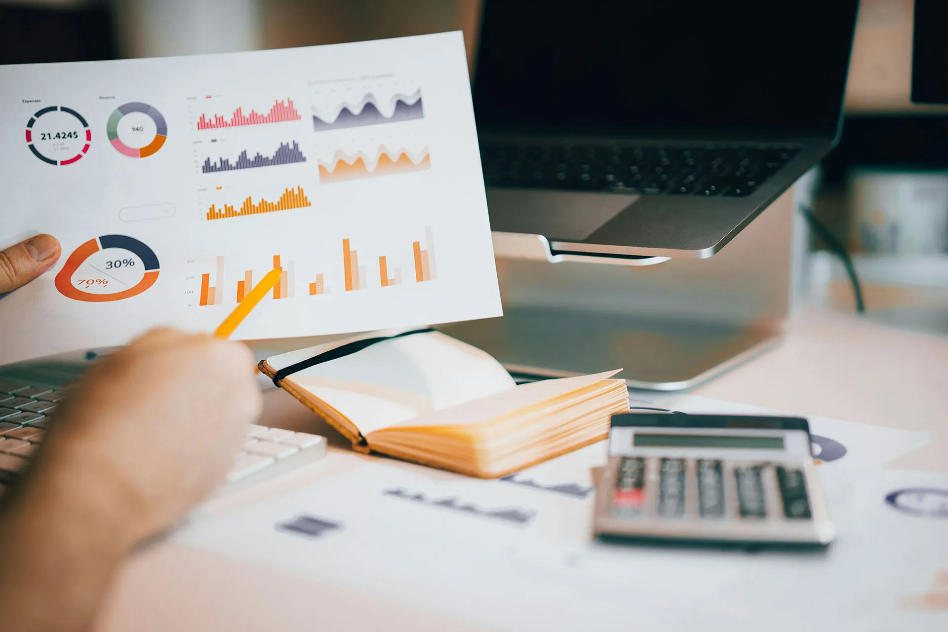 An analyst points a yellow pencil at financial charts and graphs printed on a sheet of paper at a desk featuring a calculator, laptop, and open notebook.