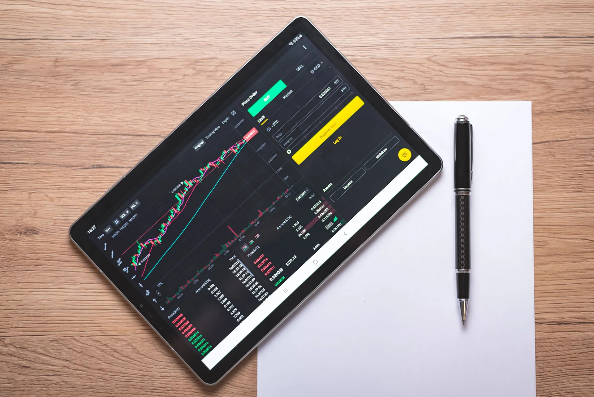  A digital display showing financial data, stock market charts, and global analytics graphics on top of a white paper and pen on a wooden table.