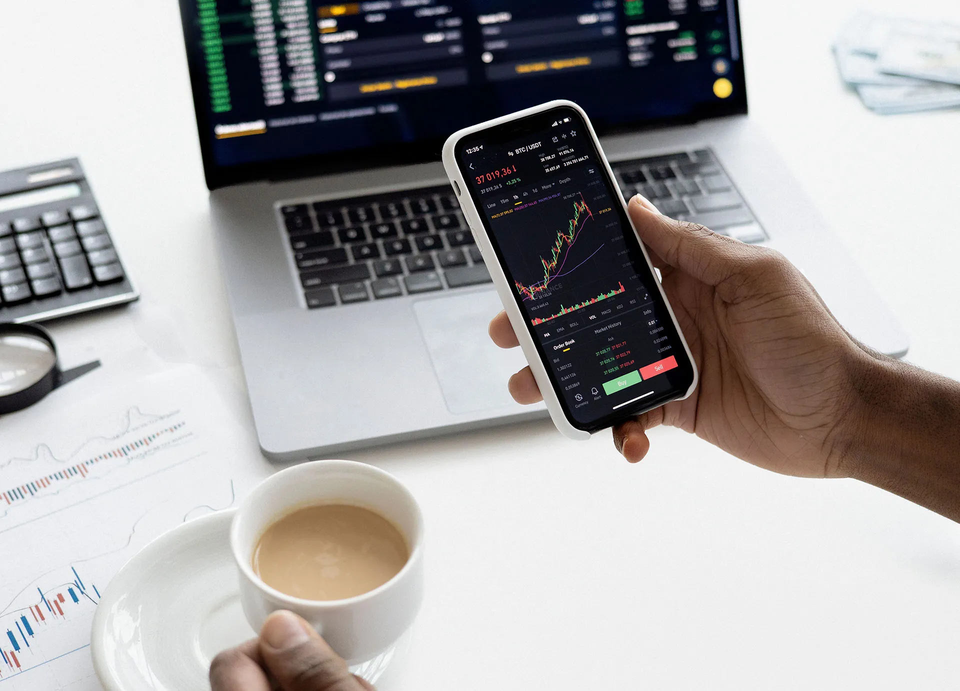 A person checking their stocks on their smartphone in front of an open laptop displaying financial statistics, while holding a cup of coffee
