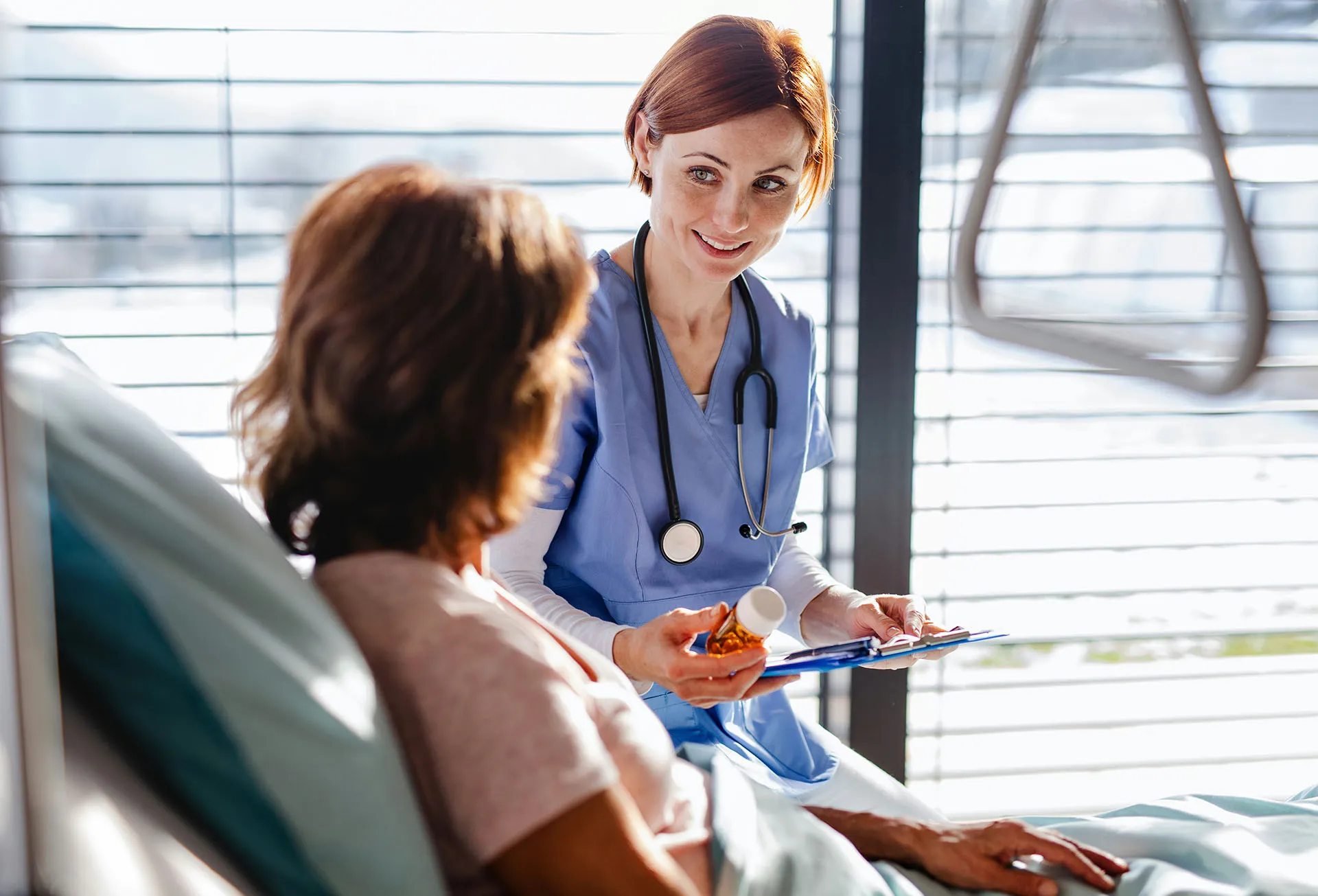  Nurse in blue scrubs discussing medication with a patient in a hospital bed while holding a clipboard.