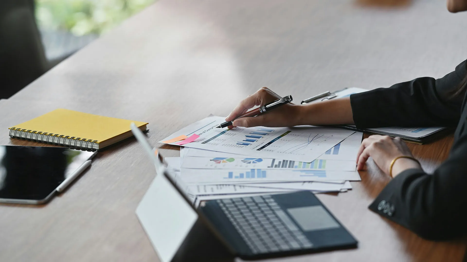 A hand holding a pen pointing to a series of printed charts, beside a notebook and laptop