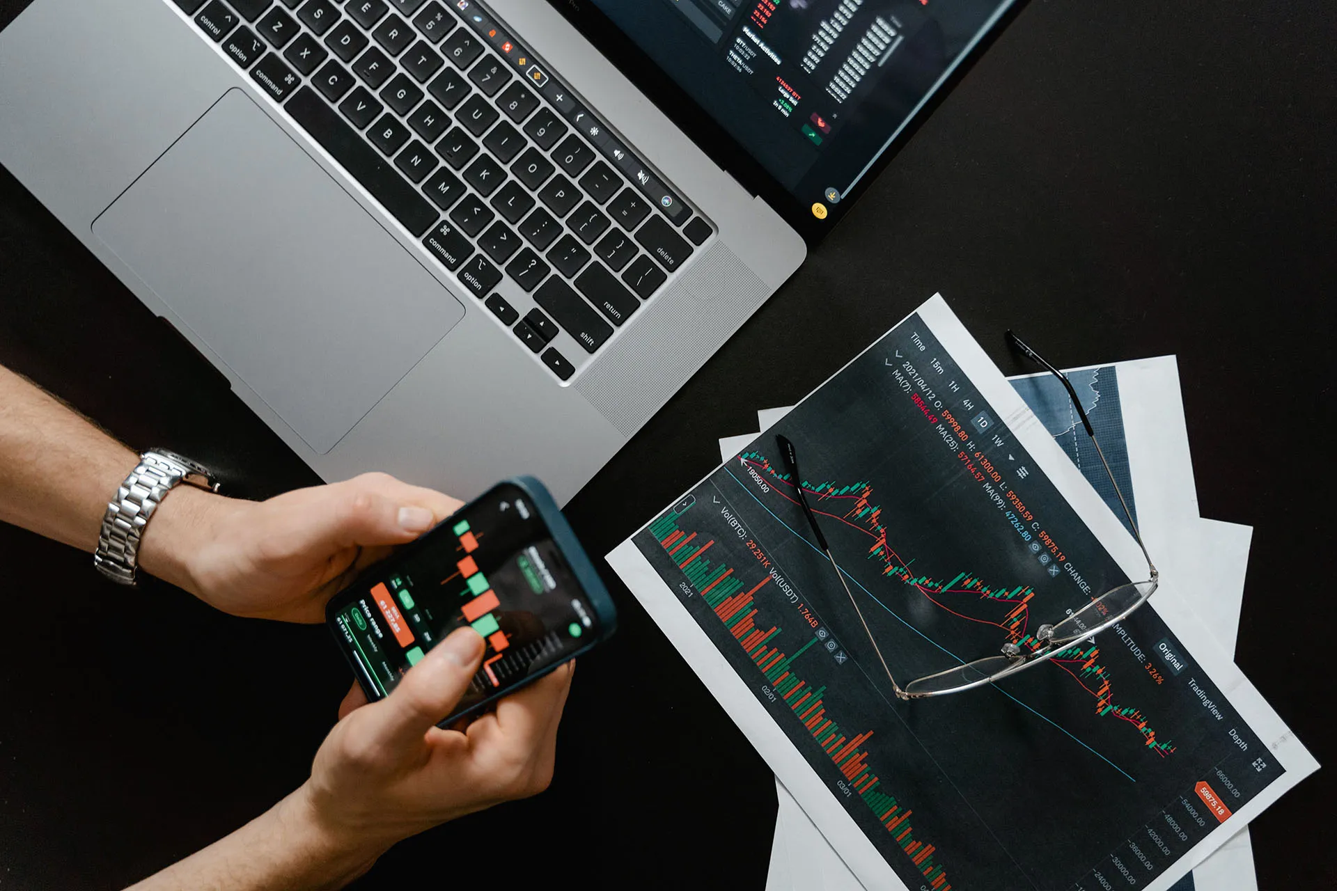 Person using a smartphone to monitor stock market charts beside a laptop, printed financial reports, and glasses on a desk.