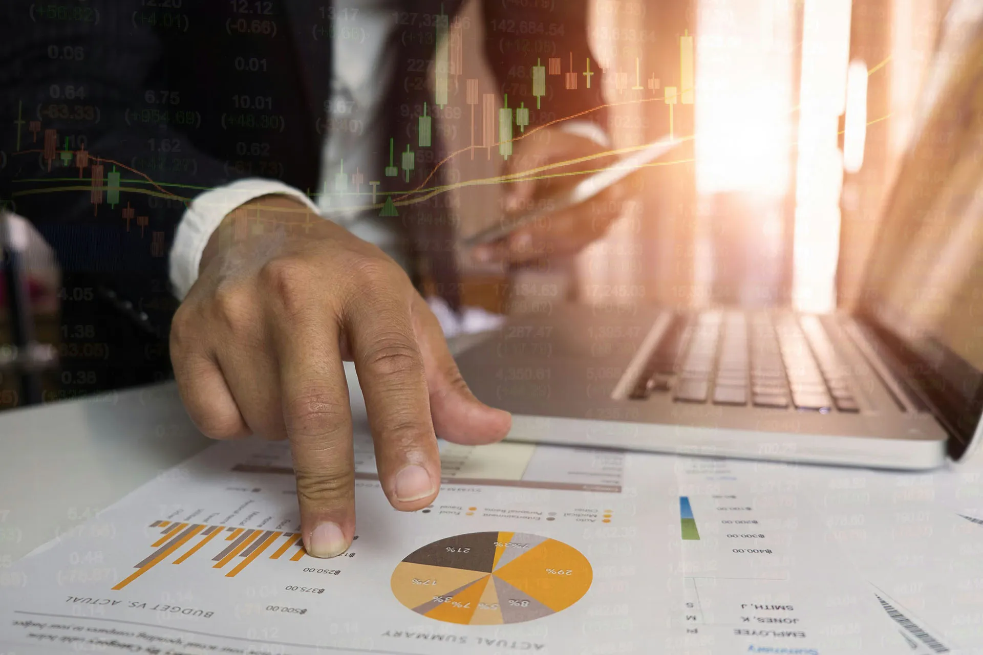  A business professional's hand pointing to printed graphs beside a laptop, with financial statistics overlaid across the image