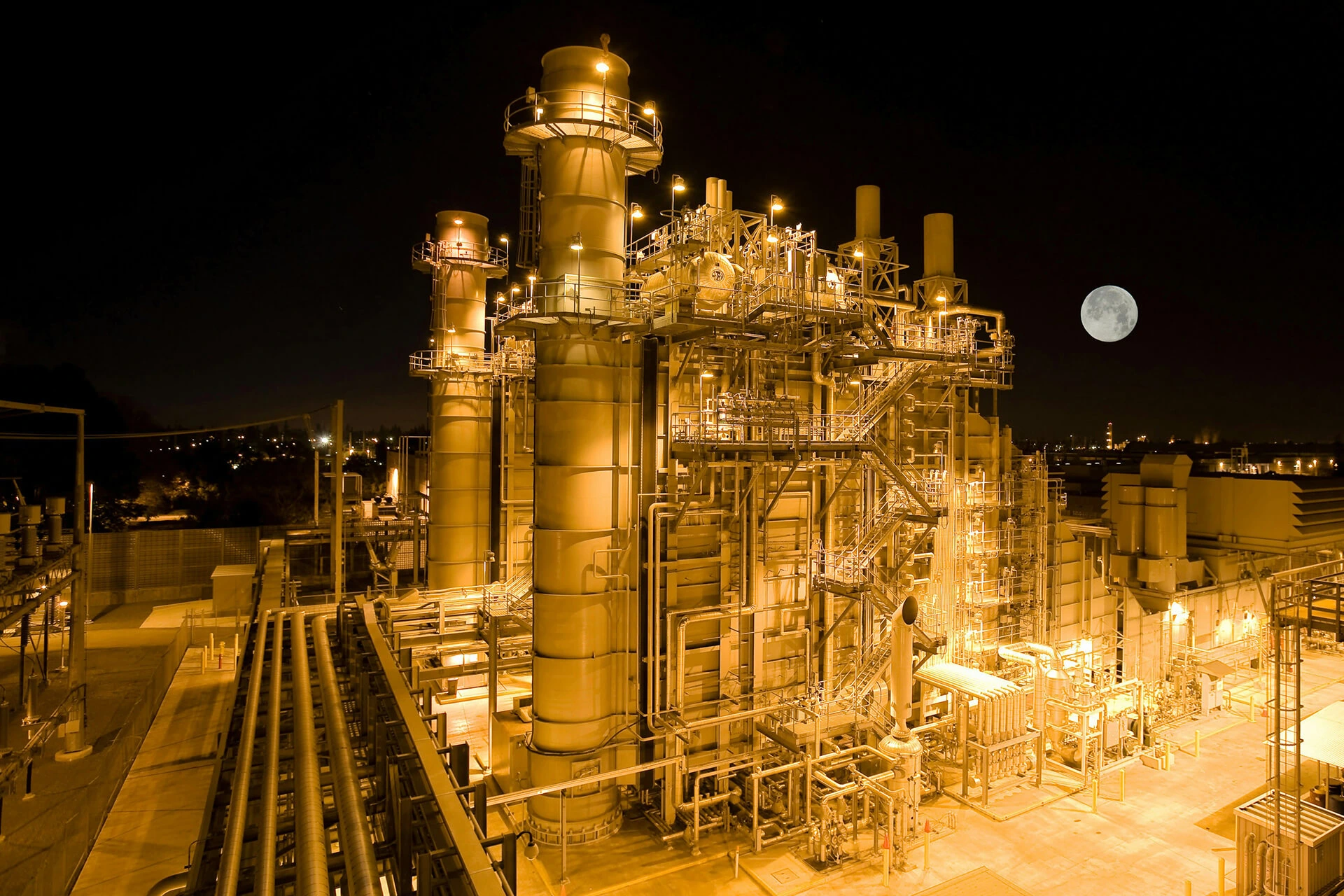 Illuminated natural gas processing facility at night with industrial pipelines, towers, and equipment under a full moon.