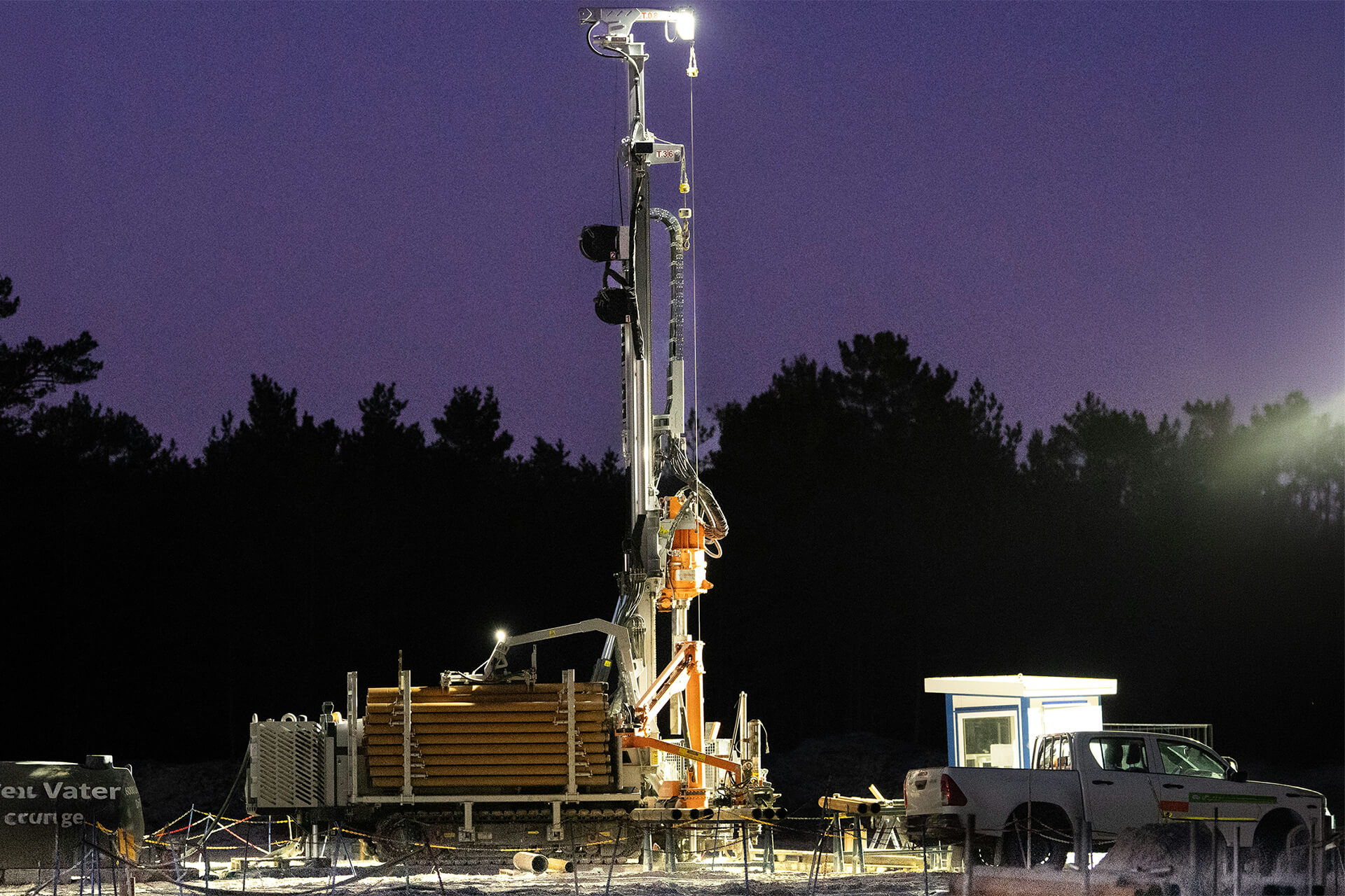 Mobile drilling rig operating at a remote energy site at night, illuminated against a dark purple sky with drilling equipment and service vehicles visible.