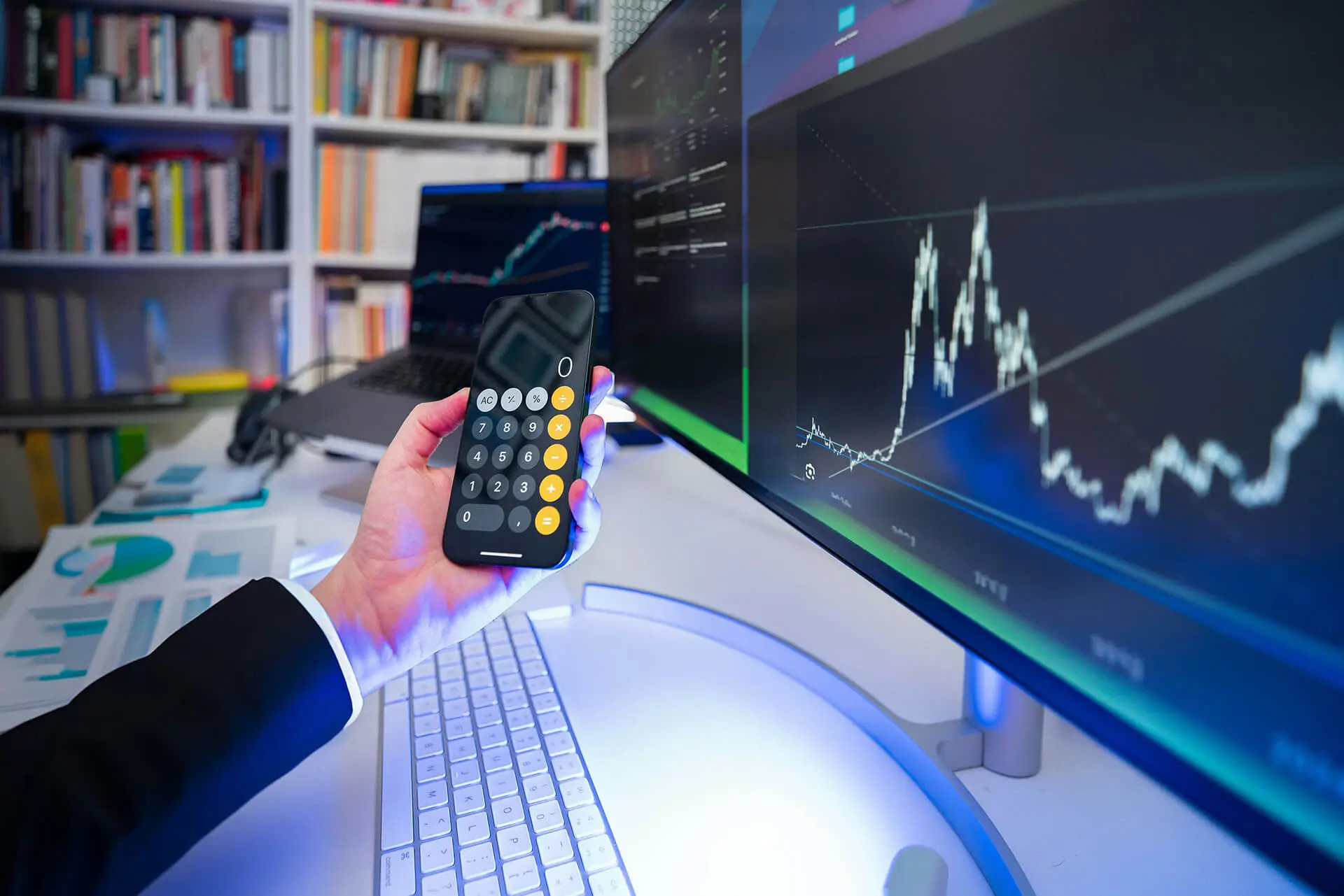 Person holding a smartphone calculator in front of multiple screens showing financial trading charts and market data.