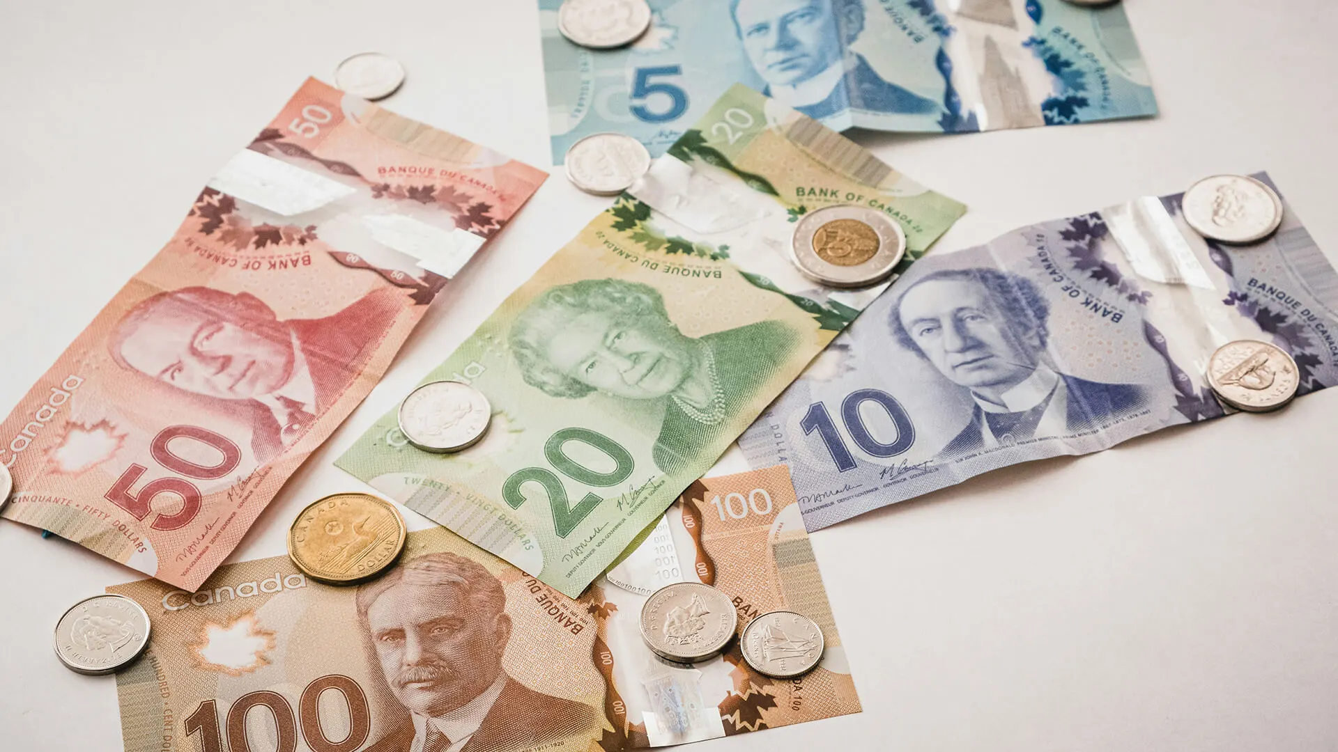 Canadian dollar bills and coins spread across a white surface, showing multiple denominations including 5, 10, 20, 50, and 100 dollar notes.