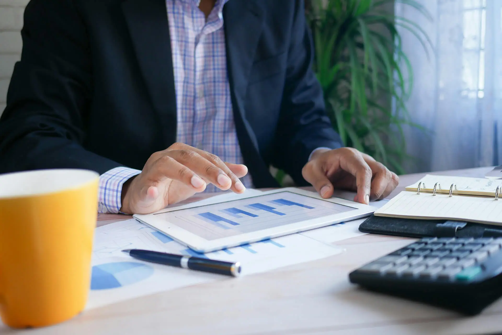 Business professional reviewing bar charts on a tablet with financial documents, a calculator, notebook, and coffee mug on the desk.